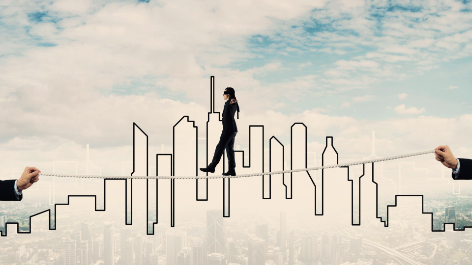 Blindfolded business person walking a tightrope with a cityscape in the background 