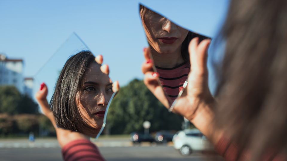Woman's hands holding two broken mirror pieces which are reflecting two parts of her face at different angles 