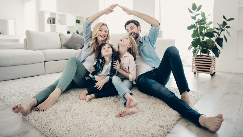Mom, dad, and two children sitting on the floor in a family room with the parents forming a roof ofver their children with their arms