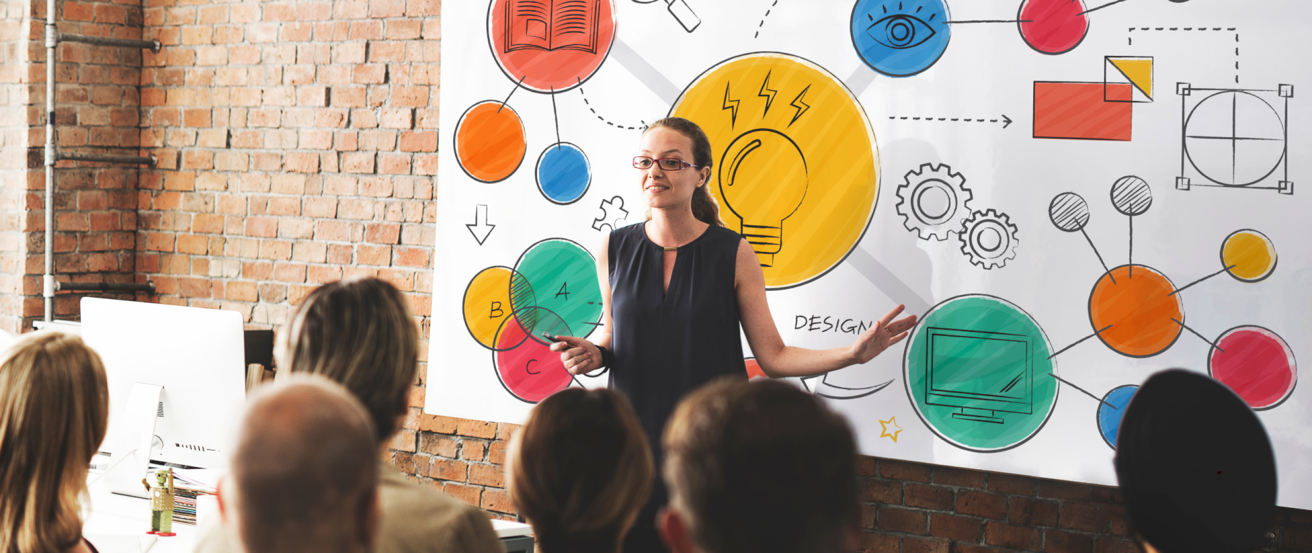 Teacher in classroom with students and display board in background showing diagram of business concepts