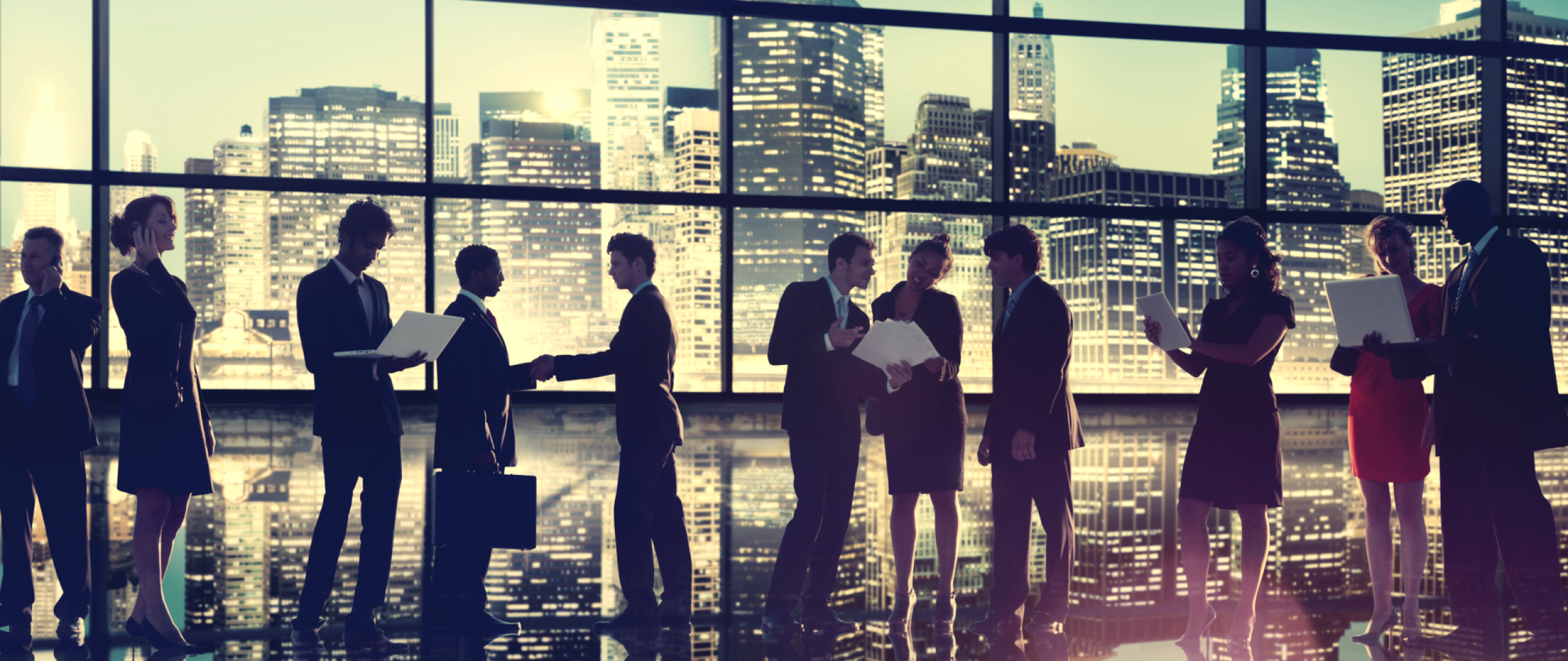 Many business people looking a papers and computers standing infornt of large window with cityscape in the background  