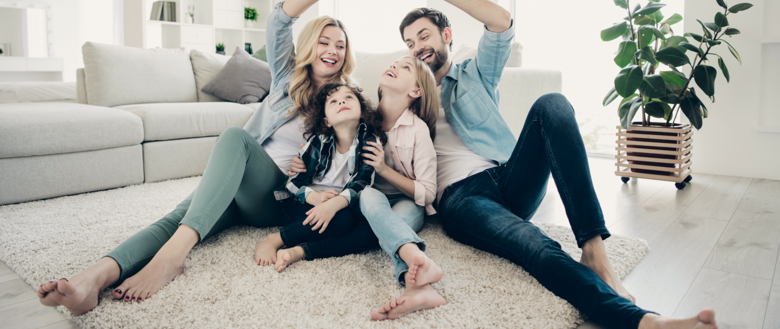 Mom, dad, and two children sitting on the floor in a family room with the parents forming a roof ofver their children with their arms
